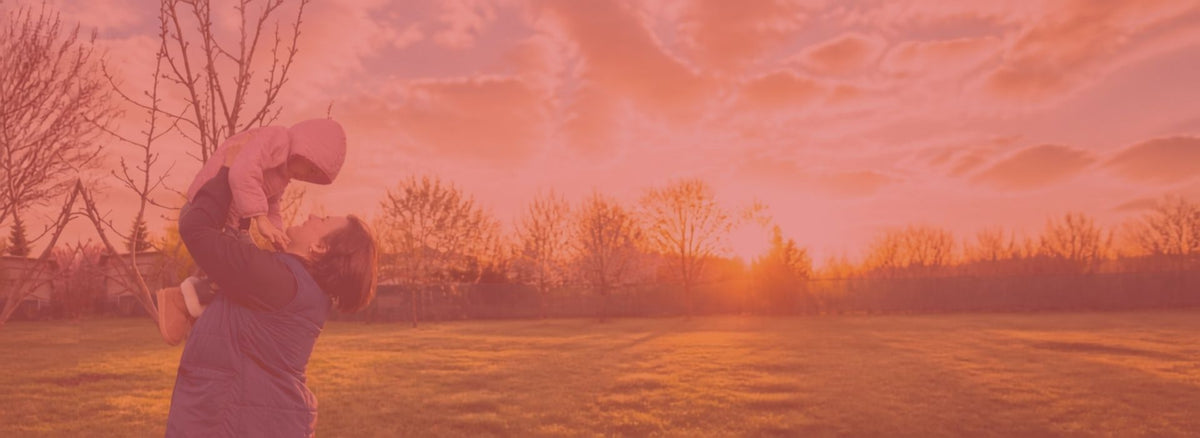 A warm-toned outdoor scene at sunset or sunrise with Geta holding a child above their head in a grassy field, under a pink-orange sky with tree silhouettes in the background.